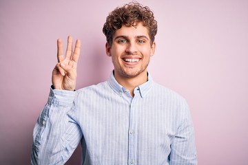 Young blond handsome man with curly hair wearing striped shirt over white background showing and pointing up with fingers number three while smiling confident and happy.