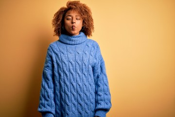 Young beautiful African American afro woman with curly hair wearing blue turtleneck sweater making fish face with lips, crazy and comical gesture. Funny expression.