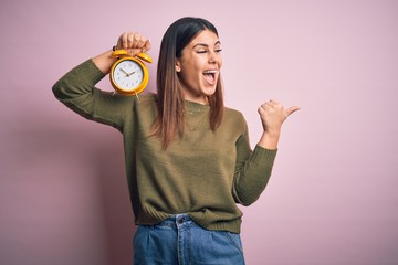 Poster - Young beautiful woman holding alarm clock standing over isolated pink background pointing and showing with thumb up to the side with happy face smiling