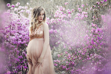 A Delicate Curly-haired Pregnant Blonde In Full Bloom Of Purple Rhododendron Flowers In A Peach Tulle Glittering Dress Hugs Her Big Tummy And Smiles, Spring Pregnancy