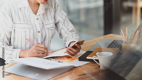 Fotografía Cropped shot of young beautiful woman in striped shirt holding smartphone in hand and writing/taking note white sitting at the modern wooden desk with modern office as background