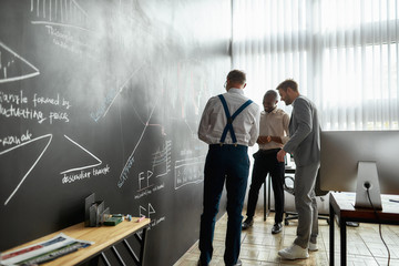 Wall Mural - One solution for business education. Full-length shot of diverse employees, traders standing near blackboard full of charts, while discussing the strategy of work. Horizontal shot