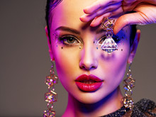 Portrait Of Beautiful Young Woman With Bright Makeup. Shiny Sequins On Face. Beautiful Brunette With Fashion Jewelry Accessories. Face Of A Girl Is Highlighted In Vivid Lights. Girl With Earrings.