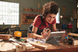 Young pretty woman with curly hair watching woodworking tutorial on digital tablet while leaning on messy table covered with sawdust
