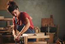 Young Concentrated Woman In Denim Overall Smoothing Just Finished Wooden Stool With Electrical Belt Sander