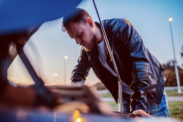 Young serious handsome caucasian bearded blond man in leather jacket looking engine of his car under the hood. Something went wrong.