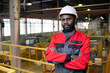 Portrait of serious confident young Afro-American factory worker in hardhat and protective suit standing in industrial shop