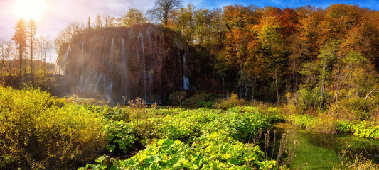 Wall Mural - Famous waterfall Prstavac in Plitvice lakes (Plitvicka jezera) national park, Croatia, large panorama. Amazing autumn sunny landscape with falling water, trees, sky and sun, outdoor travel background