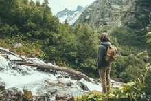 Man Traveler Alone With Backpack Looking At Mountain River Among Green Woods.