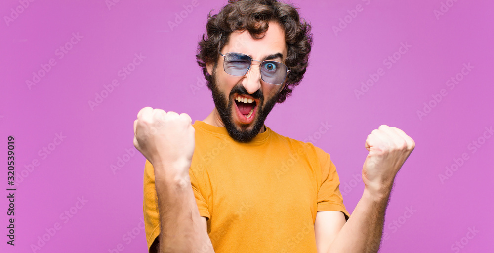young crazy cool man shouting aggressively with an angry expression or with fists clenched celebrating success against flat wall - obrazy, fototapety, plakaty 