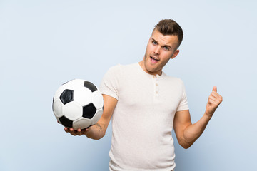 Wall Mural - Young handsome blonde man over isolated blue background holding a soccer ball