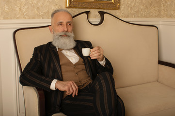 Senior handsome concentrated man with beard in business suit sitting and drinking coffee.