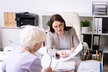 Young social worker in glasses talking to senior woman and pointing at document while assisting client in receiving services in person