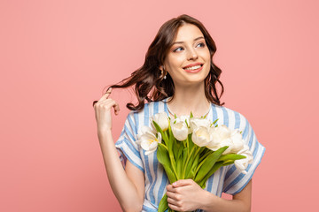 Wall Mural - happy girl touching hair while holding bouquet of white tulips and looking away isolated on pink