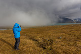 Fototapeta  - woman photographer backpack hiker at Kungsleden trail admiring nature of Sarek in Sweden Lapland with mountains, rivers and lakes, birch and spruce tree forests. Early autumn colors in stormy weather