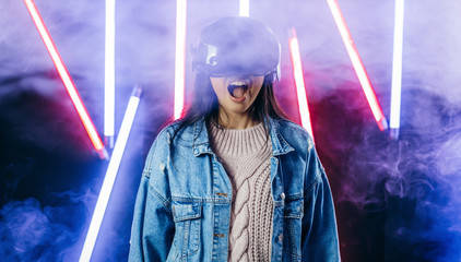 Mod curly dark haired girl dressed in blue denim jacket uses the virtual reality glasses on her head and gloves in hands in the dark studio with neon light 