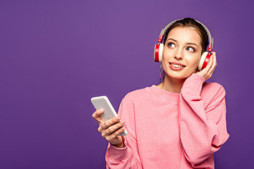 Wall Mural - smiling, dreamy girl listening music in wireless headphones while holding smartphone and looking away isolated on purple