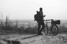 A Rider Man With A Backpack And A Bicycle Stands And Looks Into The Distance. Hilltop Overlooking A Valley In Haze, A City On The Horizon. Winter Or Autumn Landscape Road. Black And White