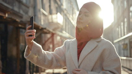 Beautiful arab woman in red hijab taking selfie in sunlight on the street. Portrait of young Muslim lady smiling looking at smartphone.