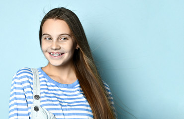 Teenage girl in jeans overall and striped sweatshirt. She is smiling while posing against blue studio background. Close up