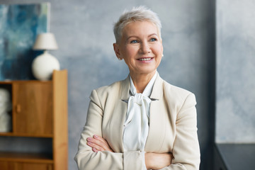 Attractive successful female professional in her fifties posing in office interior wearing stylish suit crossing arms confidently on chest and smiling. Maturity, age, people, job and occupation