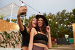 © Indico Studio/ADDICTIVE STOCK - Stylish cheerful girl friends in black hat embracing and taking selfie on mobile phone in bright day at decorated arena on festival