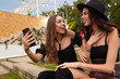 © Indico Studio/ADDICTIVE STOCK - Pleasant cheerful girl friends in black hat having fun watching photo on mobile phone sitting on green lawn near decorated stage in bright day at festival