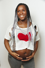 Cheerful Young Pregnant Black Female With Braids Wearing Stylish White Blouse With Red Heart Touching Belly And Looking Away With Smile While Standing Against White Wall