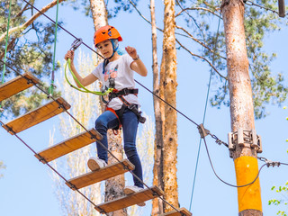 Teenager  in orange helmet climbing in trees on forest adventure park. Girl walk on rope cables and  high suspension bridge in adventure  summer city park.  Extreme sport equipment helmet and carabine