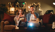 © JenkoAtaman - Family couple watching television projector at home on sofa.