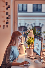 Focused blond student doing her homework while sitting next to the table and using computer.