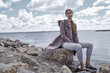 © indiraswork - Beautiful sexy young blond woman with long hair and soft natural makeup wearing jacket designer suit sitting in rock stone beach shadow ocean view water storm autumn nature fashion model.