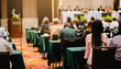 © Koonsiri - Selective focus to business woman sitting with blurry chairman of the meeting and executive committee background in auditorium for shareholders meeting or seminar event, Annual shareholder meeting.