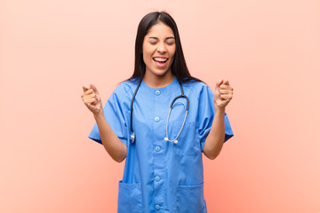 young latin nurse looking extremely happy and surprised, celebrating success, shouting and jumping against pink wall