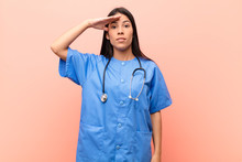 Young Latin Nurse Greeting The Camera With A Military Salute In An Act Of Honor And Patriotism, Showing Respect Against Pink Wall
