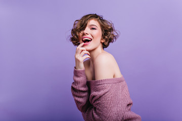 Wall Mural - Indoor portrait of sensual short-haired woman in knitted attire posing in studio. Playful curly brunette girl in sweater standing on purple background.