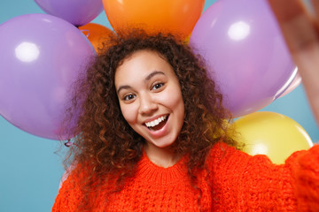 Close up of cheerful african american girl in orange knitted clothes isolated on pastel blue background with colorful air balloons. Birthday holiday party concept. Doing selfie shot on mobile phone.