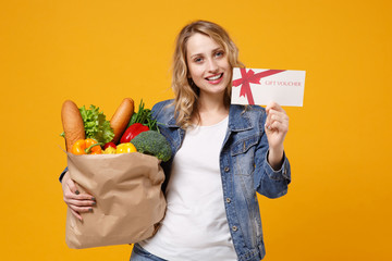 Smiling young girl in denim clothes isolated on orange background. Delivery service from shop or restaurant concept. Hold brown craft paper bag for takeaway mock up with food product gift certificate.