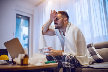 Sick caucasian unshaven man in pajamas covered with blanket sitting on sofa in living room, holding mug with tea and having fever.