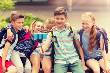 Wall Mural - primary education, technology, friendship, childhood and people concept - group of elementary school students with backpacks sitting on bench and taking picture by smartphone on selfie stick outdoors