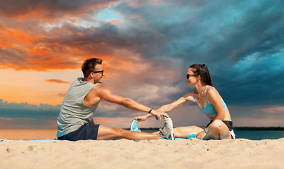 Poster - fitness, sport and lifestyle concept - smiling couple stretching legs on beach before training over sea and sunset sky on background