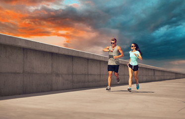 Poster - fitness, sport and lifestyle concept - happy couple in sports clothes and sunglasses running along concrete pier over sunset sky on background