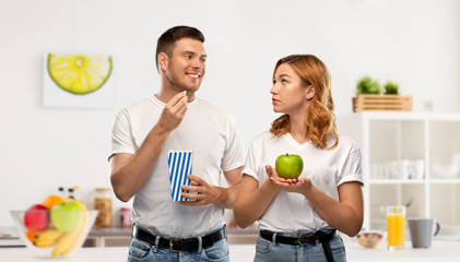 Poster - food, eating and diet concept - portrait of happy couple in white t-shirts with popcorn and green apple over home kitchen background