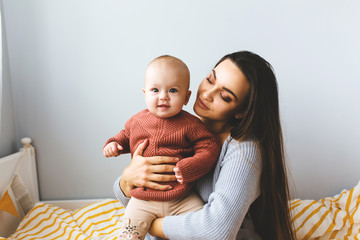 Wall Mural - A little baby girl smiles and plays on the bed with her mom, dressed in knitted sweaters pastel fashionable shades, in a room, cozy house, modern design. Happy Family at home, concept of motherhood