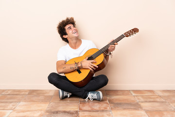 Wall Mural - Young caucasian man with a guitar sitting on the floor looking up while smiling