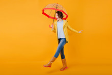 Glad Curly Girl In Jeans Funny Dancing Holding Trendy Parasol. Studio Portrait Of Inspired Young Woman In Rubber Shoes Fooling Around On Bright Yellow Background And Smiling.