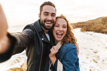 Wall Mural - image of caucasian couple taking selfie photo on cellphone by seaside
