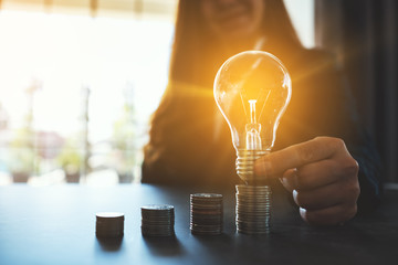 Businesswoman holding and putting lightbulb on coins stack on table for saving energy and money concept