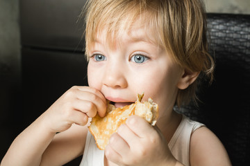 Beautiful baby with blue eyes eats delicious pancakes while sitting at kitchen table on chair.