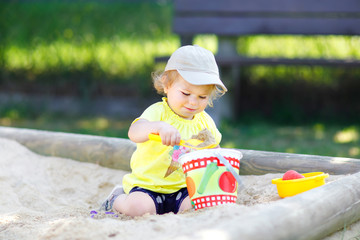 Cute toddler girl playing in sand on outdoor playground. Beautiful baby having fun on sunny warm summer sunny day. Happy healthy child with sand toys and in colorful fashion clothes.